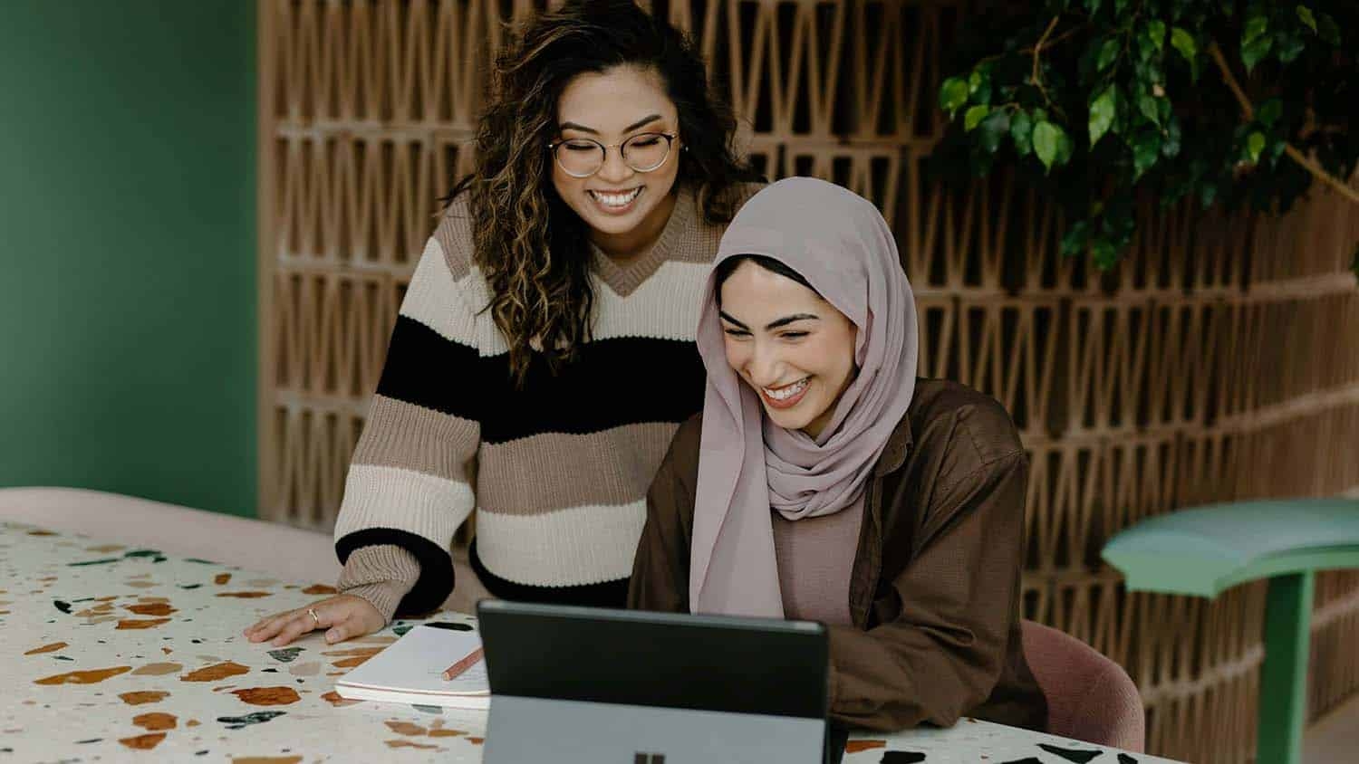 a young woman works on a computer while an older woman looks over her shoulder