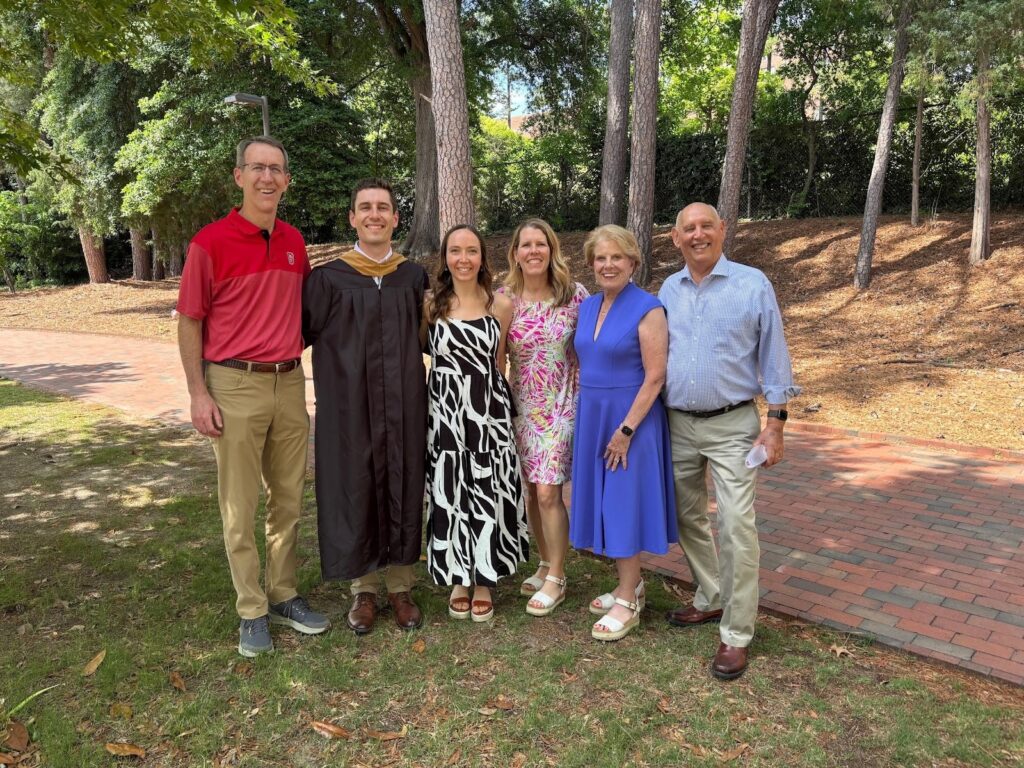 Six people posing outdoors, one in graduation attire.